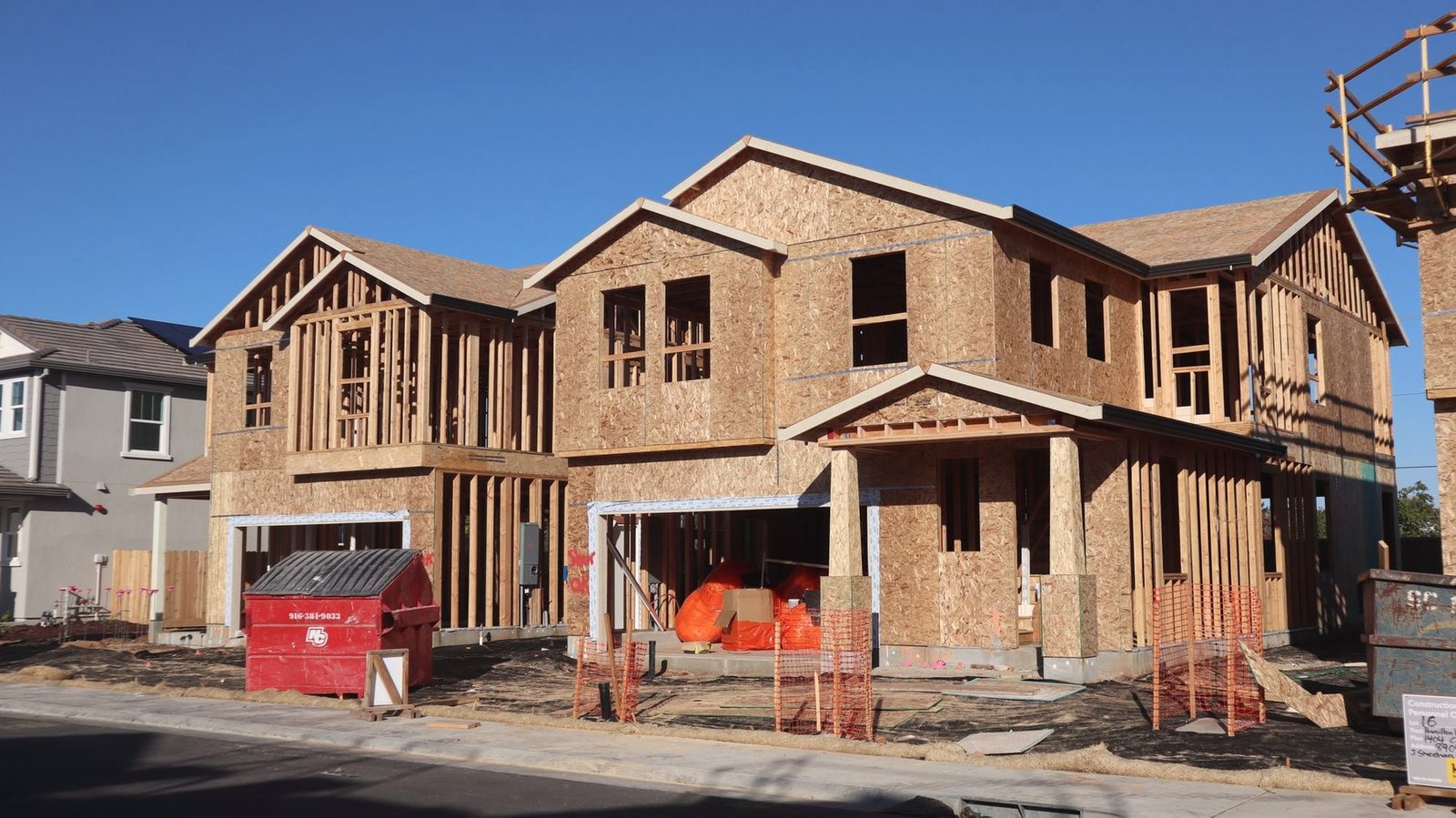 Two-story houses under construction with lumber stacks and a dumpster on the street.