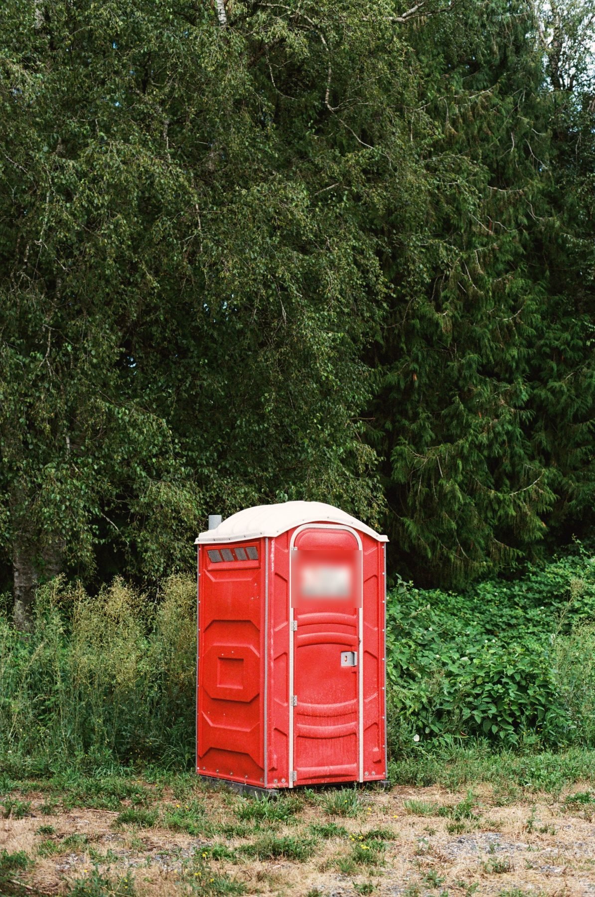 Red portable restroom unit standing alone in a grass clearing.