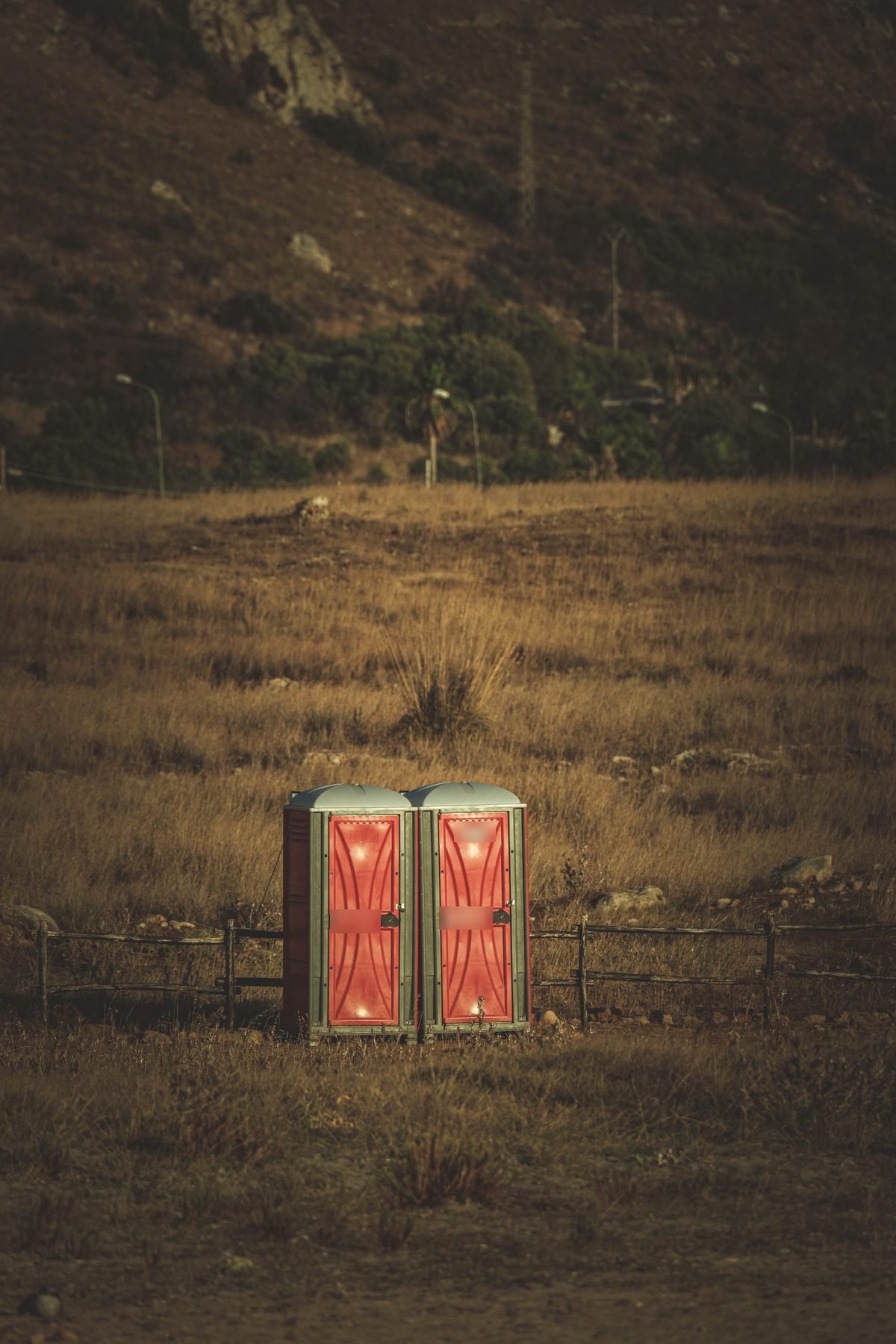Two portable restroom units standing beside a wire fence in open grassland.